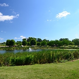 Lush green plants surround a pond with a fountain in the center at Dakotah Meadows on a bright, sunny day.